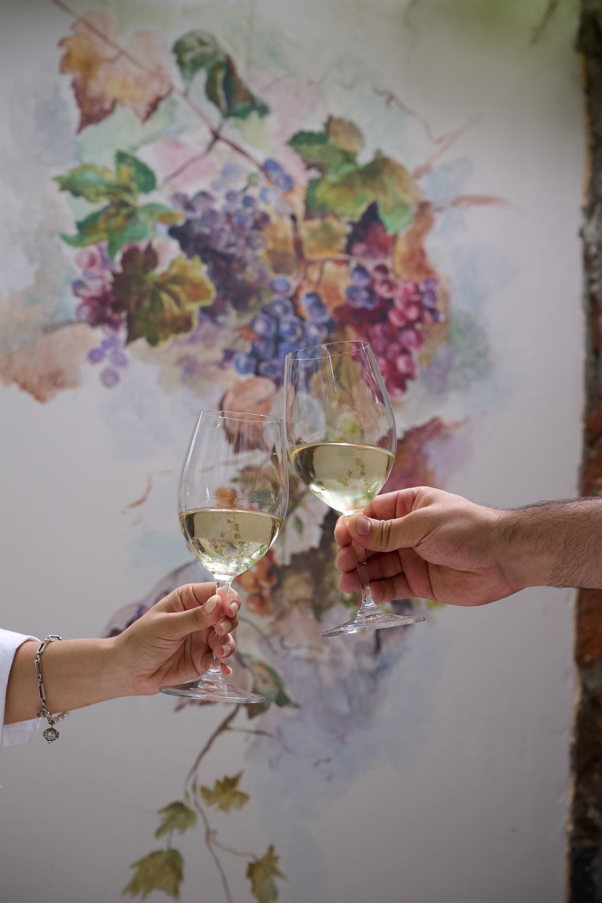 Two hands toasting with white wine before a vineyard mural